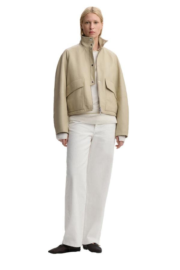 Front view of a woman wearing By Malene Birger jacket in beige with patents, zipper and pockets, paired with white jeans, white top and a pair of black flats, shown against a plain white background.